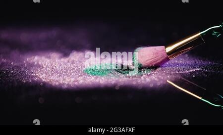 Kosmetischer Pinsel in der Nähe von rosa und türkis glänzenden Staub auf schwarzem Hintergrund Stockfoto