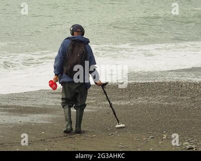 Ein Mann mit Metalldetektor läuft an einem bewölkten Tag am Strand entlang am Meer entlang Stockfoto