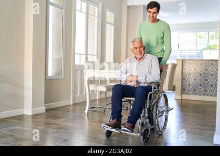 Zufriedener Senioren in einem Rollstuhl, der von seinem Sohn in der häuslichen Pflege oder in einem Altersheim betreut wird Stockfoto