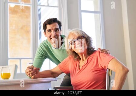 Junger Mann als Sohn, der sich in der häuslichen Pflege im Rollstuhl um seine Mutter kümmert Stockfoto