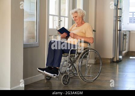 Eine zufriedene ältere Frau im Rollstuhl liest im Flur eines Pflegeheims oder Pflegeheims ein Buch Stockfoto