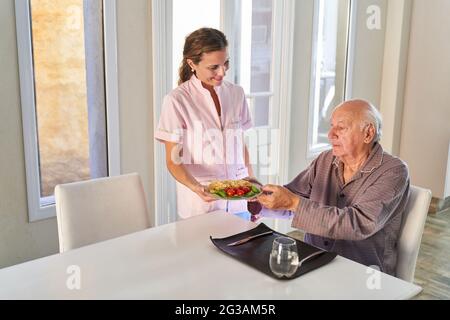 Ehefrau vom Pflegedienst oder Pflegeassistent serviert Mittagessen für einen älteren Mann in seiner Wohnung Stockfoto