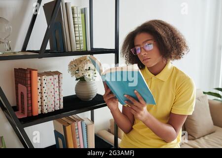 Ernst gemischtes Teenager-Mädchen mit Brille, das auf dem Sofa-Ellenbogen sitzt und in der Nähe des Regals im Wohnzimmer ein Buch liest Stockfoto