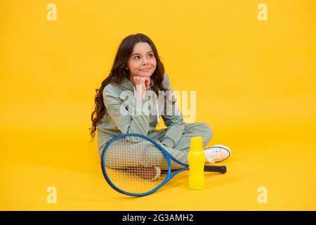 Lächelndes Kind sitzt in Sportswear mit Squash-Schläger und Wasserflasche auf gelbem Hintergrund, Sport Stockfoto