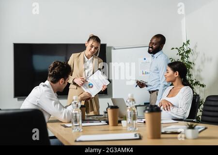 Lächelnde Geschäftsfrau, die auf Diagramme zeigt, während sie mit multiethnischen Kollegen spricht Stockfoto