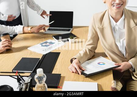 Unvollständige Ansicht einer jungen Geschäftsfrau, die in der Nähe von Infografiken lächelt, und verschwommener interrassischer Kollegen Stockfoto