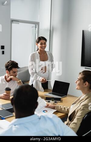 Schwanger afroamerikanische Geschäftsfrau lächelnd, während sie auf Laptop in der Nähe von Geschäftspartnern zeigt Stockfoto