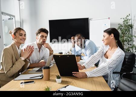 Aufgeregte multiethnische Geschäftspartner, die Clipboard in den Händen einer lächelnden afroamerikanischen Geschäftsfrau betrachten Stockfoto