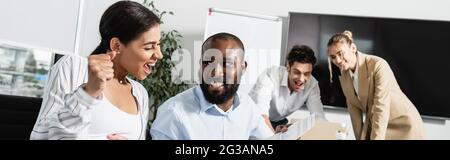 Aufgeregte afroamerikanische Geschäftsfrau mit Triumphgeste in der Nähe lächelnder Kollegen, Banner Stockfoto