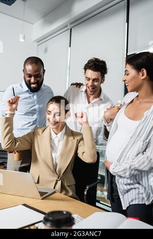 Eine aufgeregte Geschäftsfrau, die in der Nähe von Laptops und multiethnischen Kollegen eine Geste zeigt Stockfoto