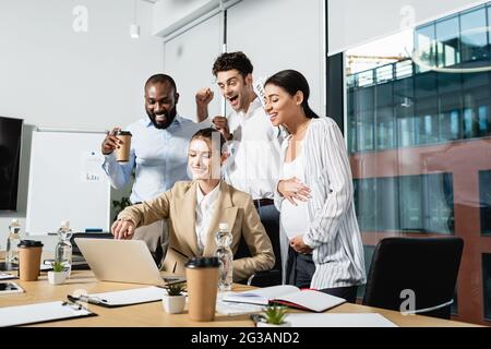 Lächelnde Geschäftsfrau, die auf den Laptop zeigt, in der Nähe aufgeregter interrassischer Kollegen Stockfoto