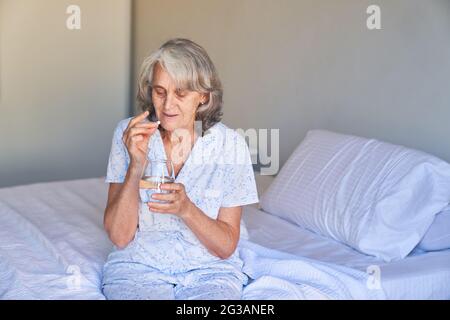 Kranke ältere Frau sitzt im Krankenhaus auf dem Bett und nimmt eine Tablette mit Wasser Stockfoto