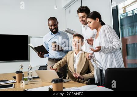 Lächelnde Geschäftsfrau, die auf den Laptop in der Nähe von glücklichen interracial Kollegen im Konferenzraum zeigt Stockfoto