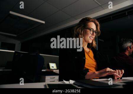 Selbstbewusste junge Geschäftsfrau, die am Arbeitsplatz sitzt, während Kollegen im Büro auf dem Laptop tippen Stockfoto