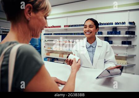 Junge glückliche Apothekerin mit Labcoat, die älteren Kunden eine Flasche Medikamente gibt, während sie ein digitales Tablet hält Stockfoto