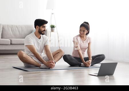 Selbstisolierung, Online-Workout, Yoga-Unterricht und Heimtraining am Morgen. Freundlicher junger arabischer Kerl und Dame, die Übungen für Beine machen und auf den Schoß schauen Stockfoto