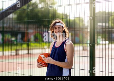 Ein hübscher junger Sportler, der in der Außenarena auf dem Smartphone im Internet surft, eine Pause vom Basketballspiel macht, die Kamera lächelt und ein mobiles Gerät benutzt. Stockfoto