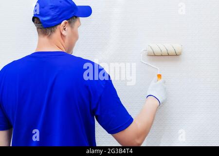 Ein Maler mit Mütze und Handschuhen bemalt die Wand mit einer Farbrolle. Porträt eines jungen Arbeiters in Uniform. Professionelle Reparatur der Räumlichkeiten. Modell Stockfoto