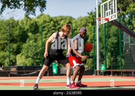 Zwei junge verschiedene Basketballspieler in Aktion in der Outdoor-Sportarena. Millennial Basketballer spielen Freundschaftsspiel zusammen, Training für Interna Stockfoto