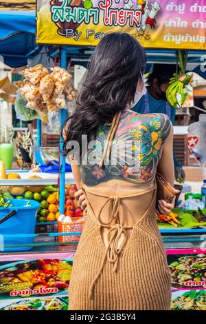 PATTAYA, THAILAND - 15. Jun 2021: Eine junge Thailänderin mit auffälliger Tätowierung auf dem Rücken bestellt für den Nachmittag eine Mahlzeit auf einem mobilen Straßenmarkt, Stockfoto