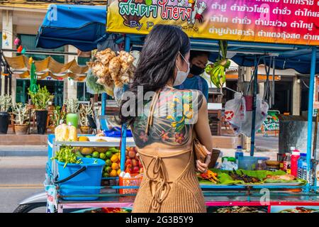 PATTAYA, THAILAND - 15. Jun 2021: Eine junge Thailänderin mit auffälliger Tätowierung auf dem Rücken bestellt für den Nachmittag eine Mahlzeit auf einem mobilen Straßenmarkt, Stockfoto