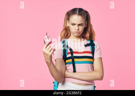 Schulmädchen mit Rucksack hält Wecker in der Hand und traurig Gesicht rosa Hintergrund Stockfoto