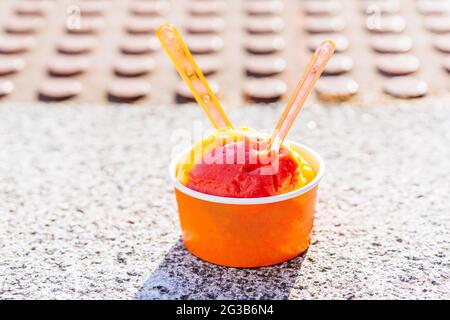 Eis aus Himbeere und Mango in einem Eisbecher zum Mitnehmen mit zwei Löffeln zum Teilen auf dem Boden auf der Straße. Konzept Eat Eis im Sommer Stockfoto