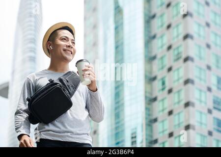Unten sehen Sie einen lächelnden jungen Vietnamesen mit Gürteltasche, der in kabellosen Ohrhörern Musik hört und eine Tasse Kaffee hält, während er mit Wolkenkratzern entlang der Straße geht Stockfoto