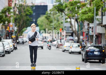 Positiver junger asiatischer Kerl in Sonnenhut, der auf einer kleinen Straßenstange gegen hohen Verkehr steht und Kaffee trinkt Stockfoto