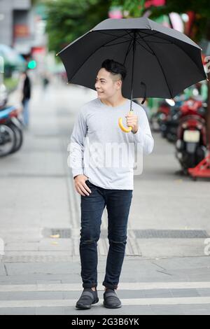 Fröhlicher junger asiatischer Kerl in legerer Kleidung, der Regenschirm hält und bei regnerischem Wetter mit Motorrollern über die Stadtstraße läuft Stockfoto