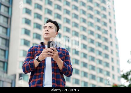 Unten sehen Sie einen ernsthaften jungen asiatischen Mann im karierten Hemd, der gegen einen Wolkenkratzer steht und die Online-Karte am Telefon verwendet Stockfoto