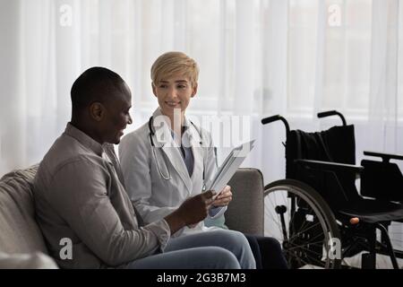 Glückliche Patienten Emotionen von positiven Testergebnissen. Lächelnde Ärztin im weißen Mantel zeigt dem erwachsenen afroamerikanischen Typ, der behindert ist, Dokumente auf der Couch wi Stockfoto