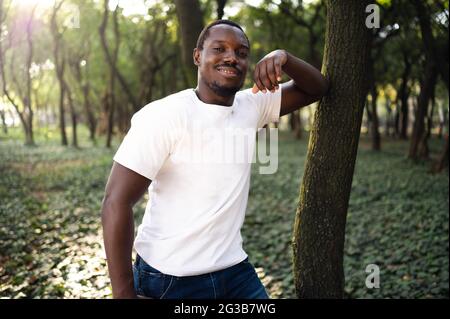 Junger lächelnder ?exikanischer Mann, der sich in einem Park auf einen Baum stützt Stockfoto