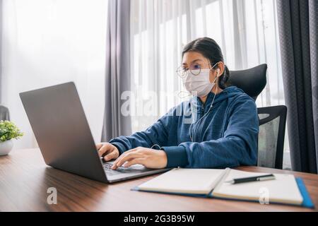 Asiatische Teenager-Mädchen tragen Kopfhörer Sprache online lernen, mit Laptop, Blick auf den Bildschirm, tun Schulaufgaben zu Hause, Notizen schreiben, zuhören Stockfoto