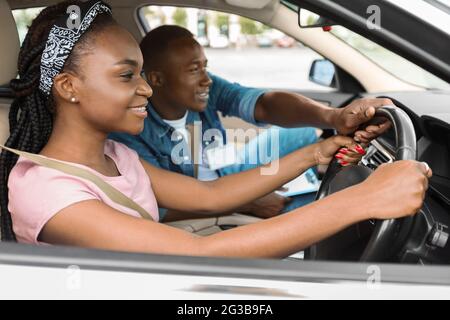 Lächelnde junge schwarze Frau, die in der Fahrschule das Parken praktiziert und mit einem hübschen Lehrer im Auto sitzt. Fröhlicher afroamerikanischer Mann, der Coach fährt Stockfoto