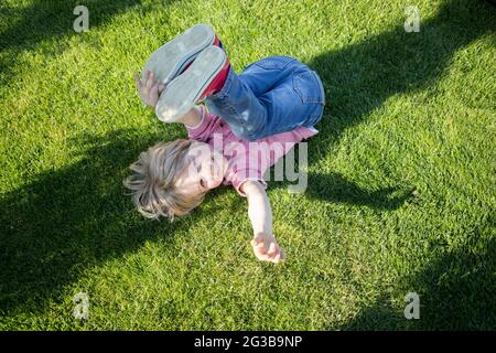 Glücklicher kleiner Junge liegt kopfüber auf dem grünen Gras im Park. Kinderglück, Freude, Selbstgenuss. Gute Laune, entspannen Stockfoto