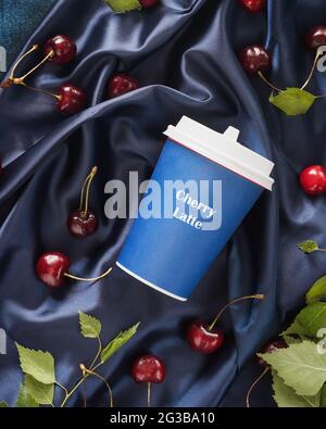 Cherry Latte, eine neue Art von Kaffee. Lebensmittelkomposition mit einer Tasse Kaffee, Kirschen und Blättern auf einem blauen, satinierten, gefalteten Hintergrund. Kaffee zum trinken Stockfoto
