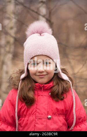 Portrait Von Happy Little Girl In Leuchtend Roten Jacke Und Gestrickter Rosa Kappe Auf Dem Hintergrund Des Frühlings Im Freien. Stockfoto