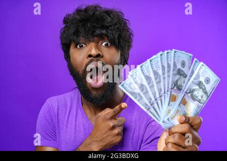 Schwarzer aufgeregt Mann hält Haufen von Dollar-Banknoten vor purpurem Hintergrund Stockfoto