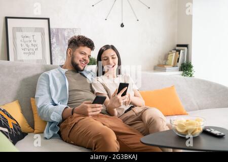 Fröhliches Paar mit Smartphones, während sie sich auf der Couch im Wohnzimmer ausruhen Stockfoto