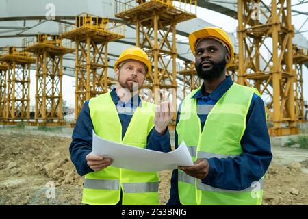 Bärtiger männlicher Ingenieur mit Blaupause, der einem afrikanischen Kollegen einen Neubau zeigt Stockfoto