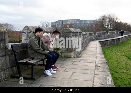 Zwei chinesische Staatsbürger (einer trägt eine Gesichtsmaske) machen eine Verschnaufpause auf den Stadtmauern von York, hinter denen sich das Staycity Hotel aufragt, wo die Tannen waren Stockfoto