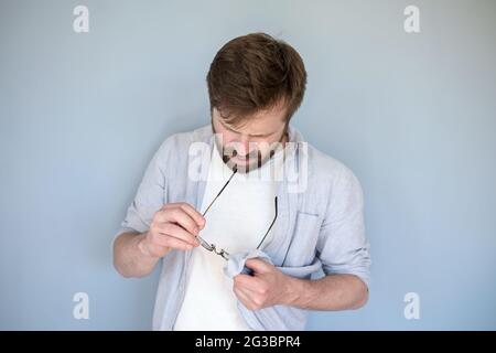 Konzentrierter kaukasischer Mann reinigt Gläser von Staub und Schmutz und wischt sie mit seinem Hemd ab. Grauer Hintergrund. Stockfoto