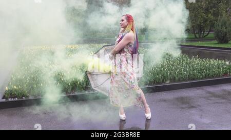 Entzückendes Mädchen in einem Kleid mit einem Blumendruck mit Regenbogen Zöpfen und Make-up. Tanzt mit einem Regenschirm, der sich in gelbem Rauch vor der Kulisse von FL Stockfoto