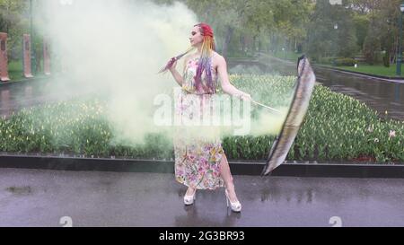 Entzückendes Mädchen in einem Kleid mit einem Blumendruck mit Regenbogen Zöpfen und Make-up. Tanzt mit einem Regenschirm, der sich in gelbem Rauch vor der Kulisse von FL Stockfoto