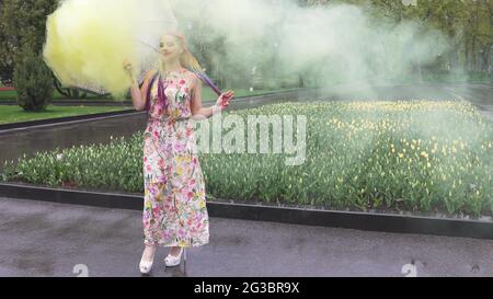 Entzückendes Mädchen in einem Kleid mit einem Blumendruck mit Regenbogen Zöpfen und Make-up. Tanzt mit einem Regenschirm, der sich in gelbem Rauch vor der Kulisse von FL Stockfoto