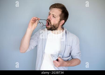 Der junge bärtige Kaukasusmann reinigt seine Brille von Staub und Schmutz, atmet auf die Linsen und wischt sie mit Hemd ab. Stockfoto