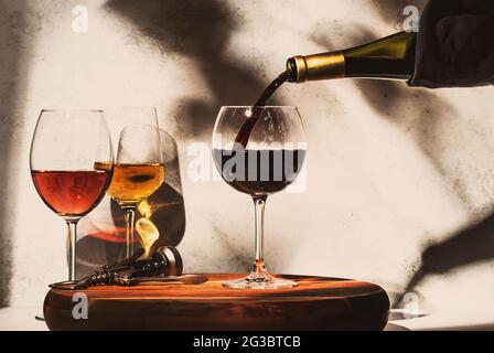 Weinprobe. Rotwein, der auf dem Hintergrund in Glas gegossen wird, mit einer Auswahl an Rot-, Weiß- und Roséweinen in Gläsern. Hartes Sonnenlicht und Schatten vom Laub Stockfoto