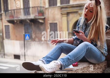 Trendige junge blonde Frau, die raucht, während sie ihr Mobiltelefon im Freien in der Stadt benutzt, während sie an einer Wand in der Altstadt sitzt Stockfoto
