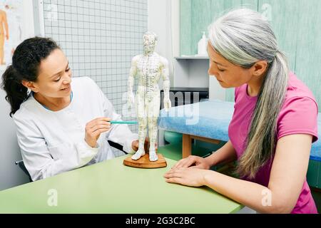 Weibliche Akupunkturistin zeigt ihrem Patienten im Zentrum für traditionelle chinesische Medizin Punkte zum Akupunkturmodell des menschlichen Körpers Stockfoto